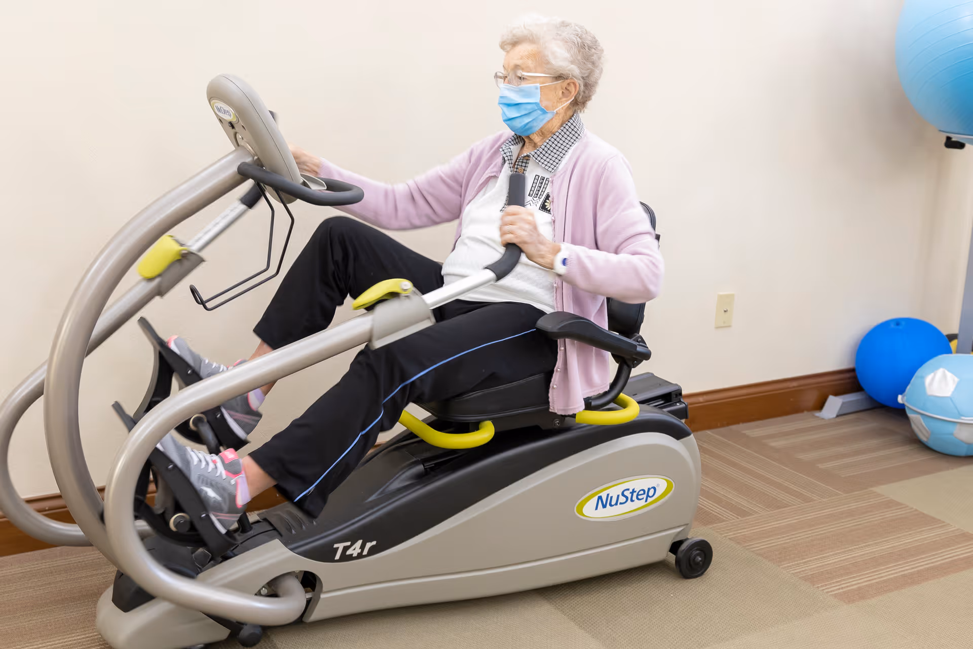 An elderly woman wearing a face mask and glasses exercises on a NuStep T4r recumbent cross trainer machine in a room with beige walls and carpeted floor. There are blue exercise balls in the background.