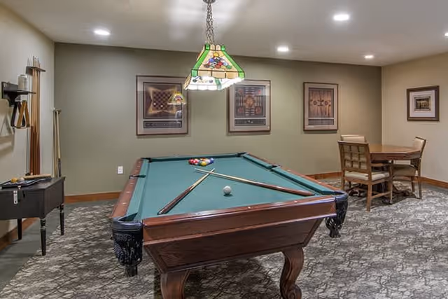 A recreational room with a pool table in the center, pool cues resting on the table, and a stained glass hanging light fixture above. The room has patterned carpet, framed artwork on the walls, and a small table with chairs in the corner.