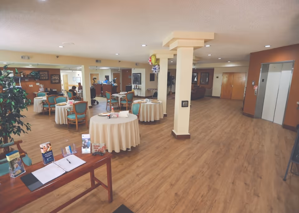 Large communal area in a senior living facility with round tables and chairs, a reception desk and elevators.