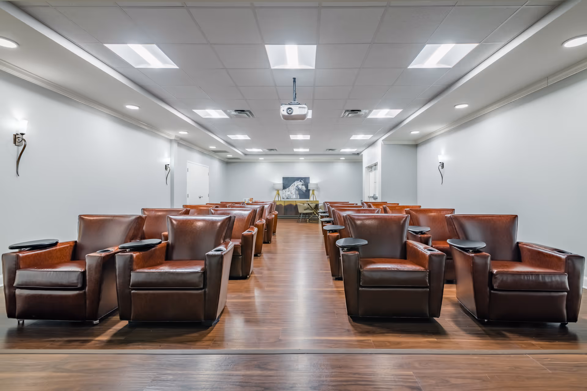 A room with rows of brown leather armchairs equipped with small black trays, arranged facing a wall with a painting of a white horse and two lamps on either side. The room has a wooden floor, white walls, and a ceiling with multiple recessed lights and a ceiling-mounted projector.