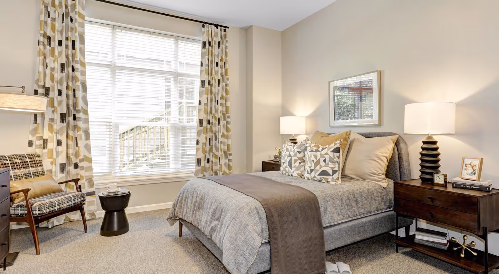 Well-lit bedroom with a made bed, two nightstands and lamps, patterned curtains, and an armchair by a large window.