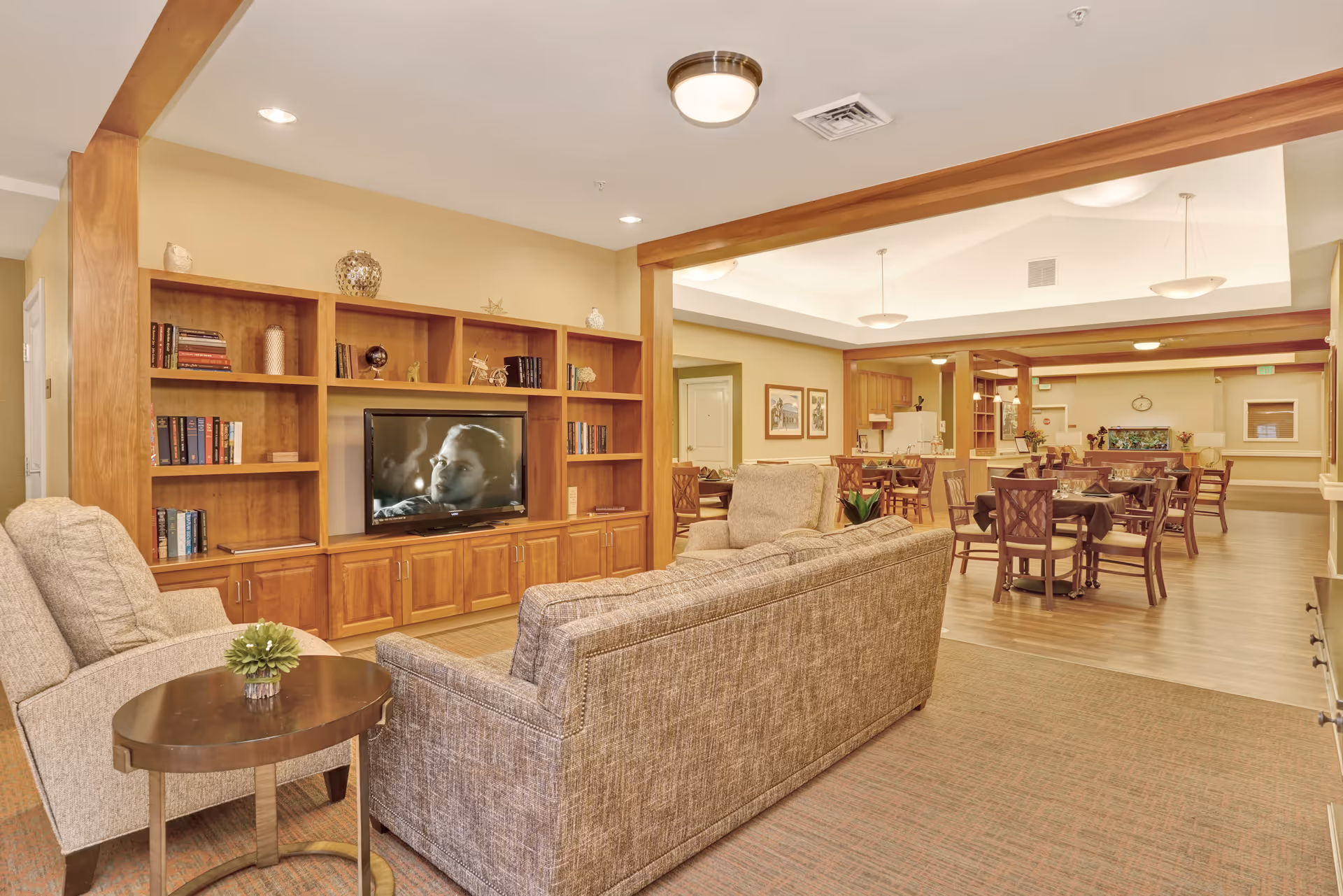 A spacious and well-lit common area in a senior living facility featuring a comfortable seating arrangement with a sofa and armchair facing a wooden entertainment center with a television. The background shows a dining area with several tables and chairs, warm beige walls, and ceiling lights.