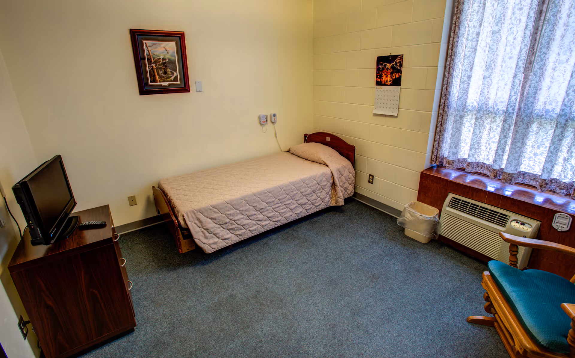 A small bedroom with a single bed covered with a beige quilt, a wooden dresser with a TV and remote on top, a framed picture on the wall, a calendar hanging on the wall, a window with lace curtains, an air conditioning unit below the window, a trash can, and a wooden rocking chair with a green cushion.
