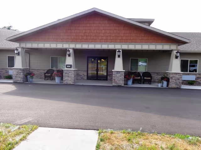 Front exterior view of a single-story assisted living facility with a covered entrance supported by stone pillars. There are chairs and potted plants arranged on the porch area near the entrance doors.