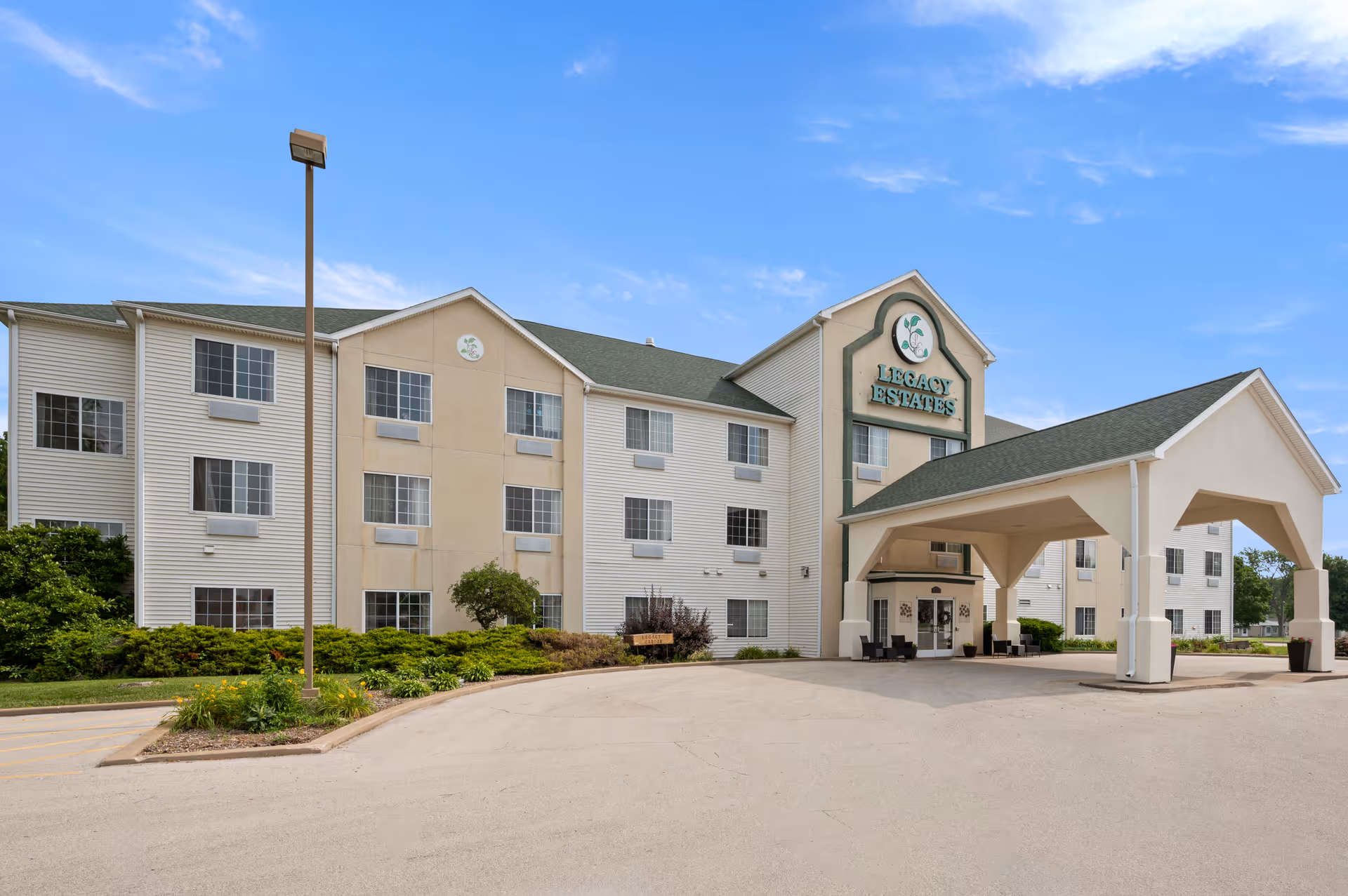 Exterior view of Legacy Estates of Monmouth, a three-story senior living facility with a covered entrance driveway, surrounded by greenery and a clear blue sky.