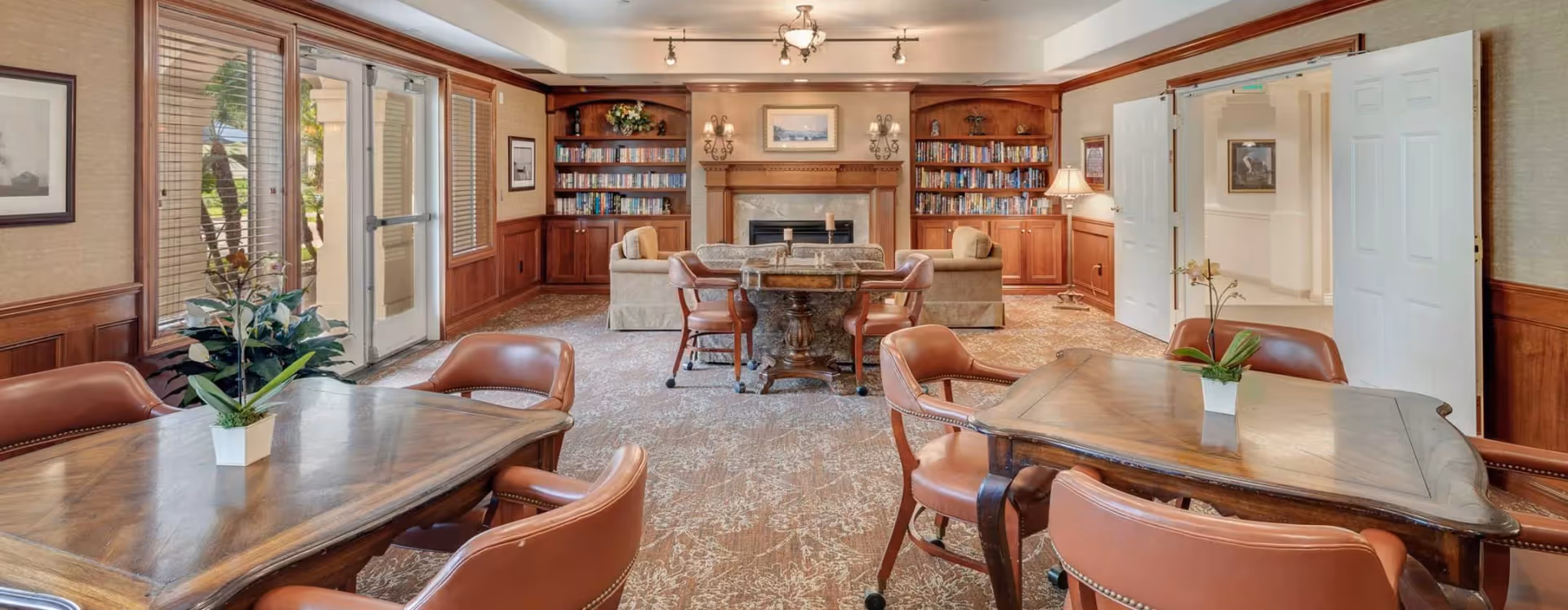 Communal lounge with wooden tables and leather chairs, a fireplace flanked by bookshelves, and glass doors leading outside.