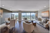 A spacious and modern living room with large windows offering a scenic view. The room features a white sofa with cushions, two beige armchairs, a round wooden coffee table, a patterned rug, and a dining area with a glass table and four chairs. The walls are decorated with artwork, and the floor is dark wood.
