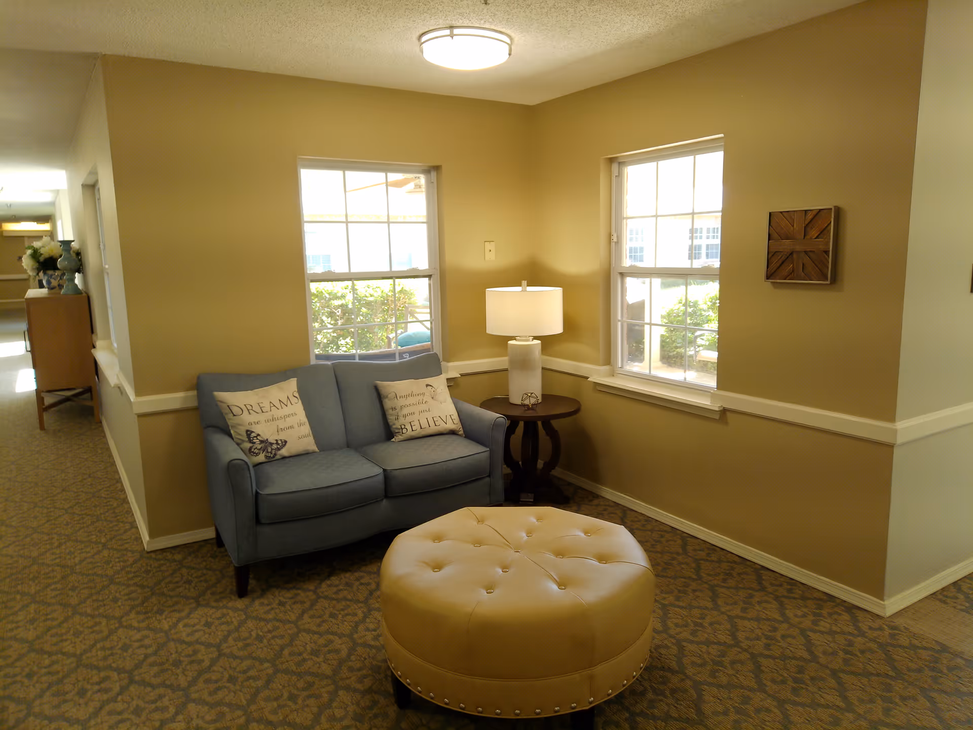 A cozy corner seating area in a senior living facility with a blue loveseat adorned with two decorative pillows, a round beige tufted ottoman, and a small wooden side table with a white lamp. Two windows let in natural light, and the walls are painted a warm beige color. The carpet has a patterned design, and a hallway with furniture and flowers is visible in the background.