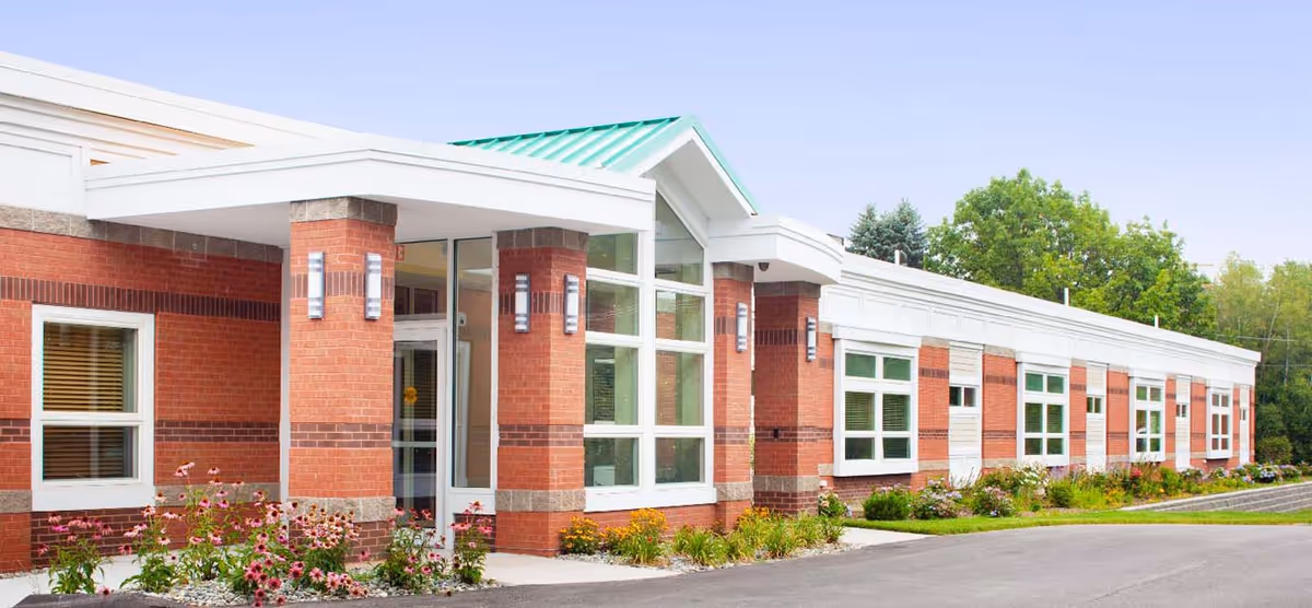 Exterior view of a single-story brick building with large windows and a green metal roof over the entrance. There are flower beds with pink and yellow flowers along the front of the building and trees in the background under a clear sky.