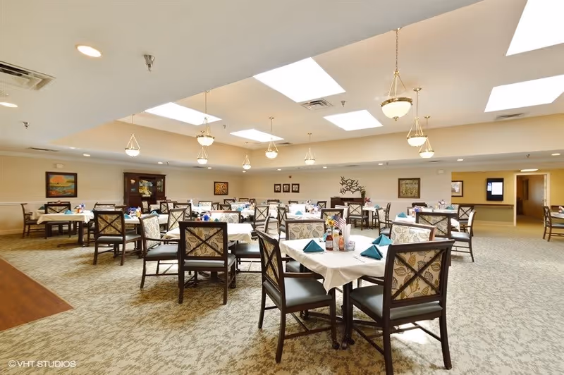 A spacious dining room with multiple tables covered with white tablecloths and set with teal napkins, condiments, and small flower arrangements. The room has patterned carpet, beige walls adorned with framed artwork, and ceiling lights with hanging fixtures and skylights providing natural light.
