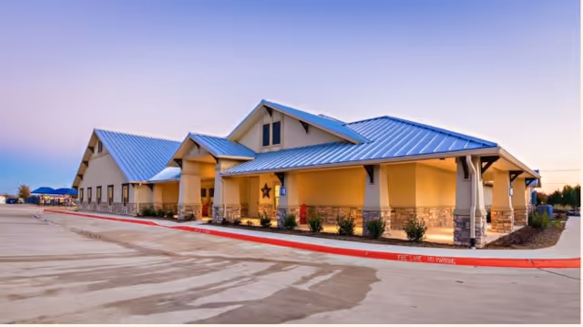 Exterior view of a single-story building with a metal roof and beige walls, featuring a covered entrance and small shrubs along the foundation. The building is situated next to a large paved area with a red curb and clear sky at dusk.