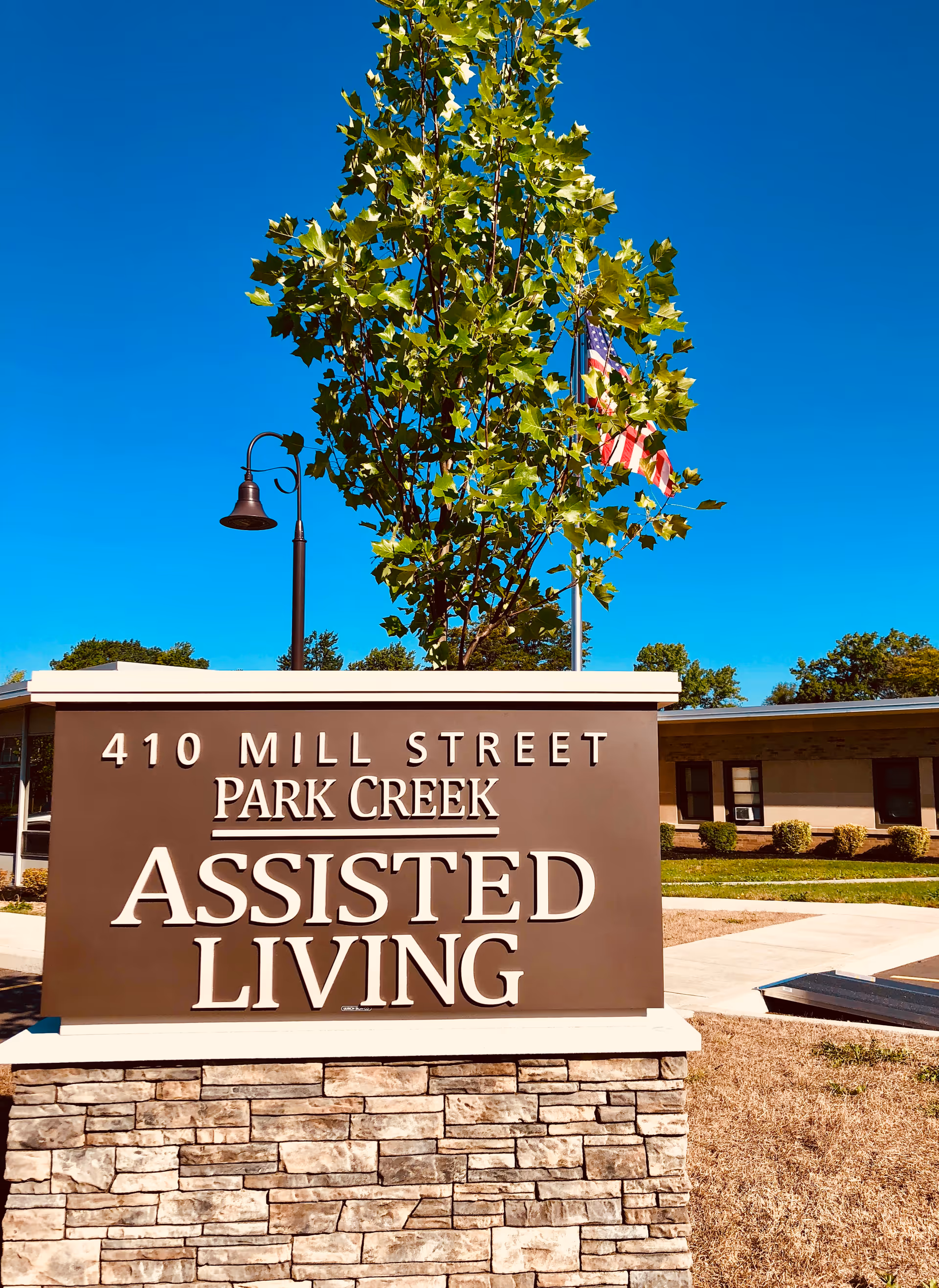 Outdoor view of a sign for Park Creek Assisted Living at 410 Mill Street with a tree and an American flag in the background under a clear blue sky.