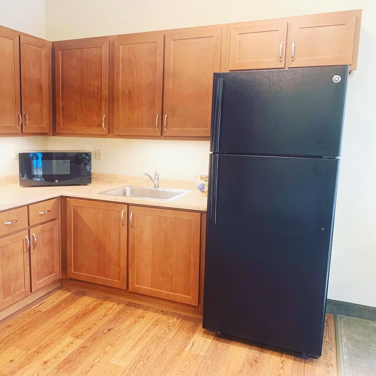 A kitchen area with wooden cabinets, a black refrigerator, a microwave on the countertop, a stainless steel sink, and a wooden floor.