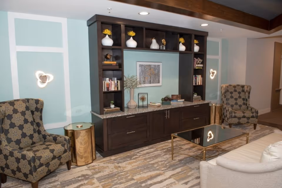 Stylish common living area with two patterned armchairs, a dark wood built-in cabinet and shelves, a glass-topped coffee table, and decorative accents.