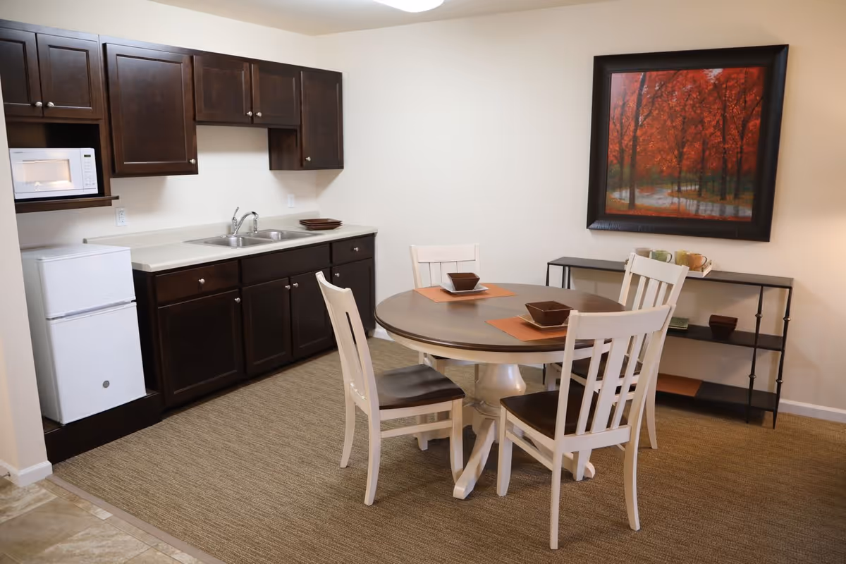 A small dining area with a round wooden table and four white chairs with dark seats. The table is set with placemats and square bowls. Behind the table is a black metal shelf with cups and bowls. To the left is a kitchenette with dark wooden cabinets, a white mini refrigerator, a microwave, and a double sink. A framed painting of red autumn trees hangs on the wall above the shelf.
