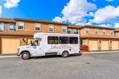 A white shuttle bus with the name Ponté Palmero parked in front of a row of tan-colored townhouses under a blue sky with scattered clouds.
