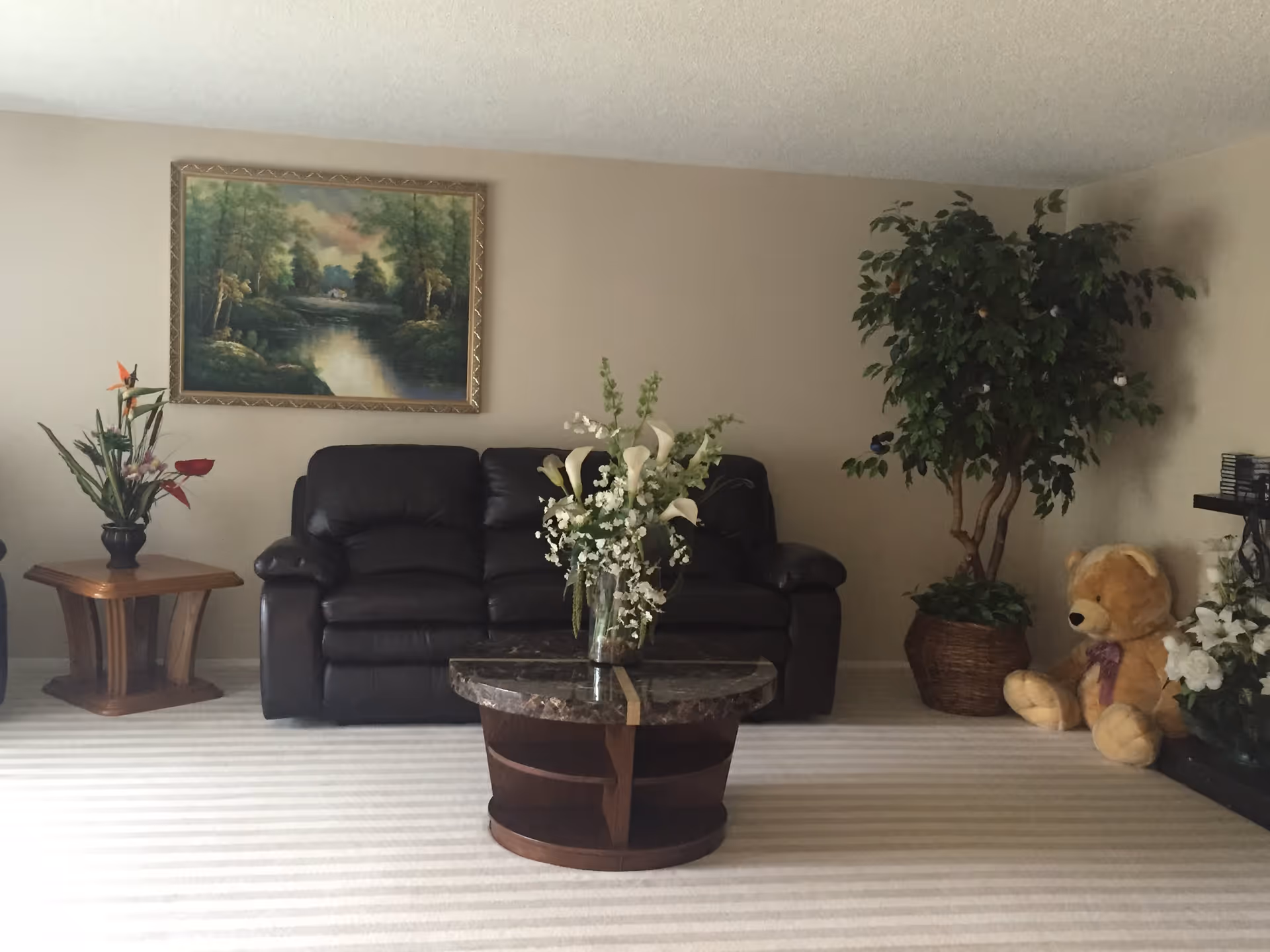 A cozy living room with a dark leather sofa against a beige wall, a round wooden coffee table with a floral arrangement in the center, a side table with a vase of flowers on the left, a large potted plant and a teddy bear on the right, and a framed landscape painting hanging above the sofa.