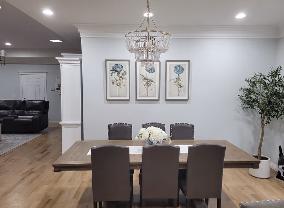 A dining room with a wooden table and six gray upholstered chairs. A white runner and a floral centerpiece are on the table. Three framed botanical prints hang on the wall above the table. A chandelier with crystal elements hangs from the ceiling. To the right, there is a potted plant, and in the background, a living room area with a black leather sofa is partially visible.