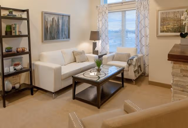 A cozy living room with a white sofa, a white armchair with a throw blanket, a glass-top coffee table with a small plant, a floor lamp, a bookshelf with decorative items, and framed artwork on the walls. There is a window with patterned curtains letting in natural light.
