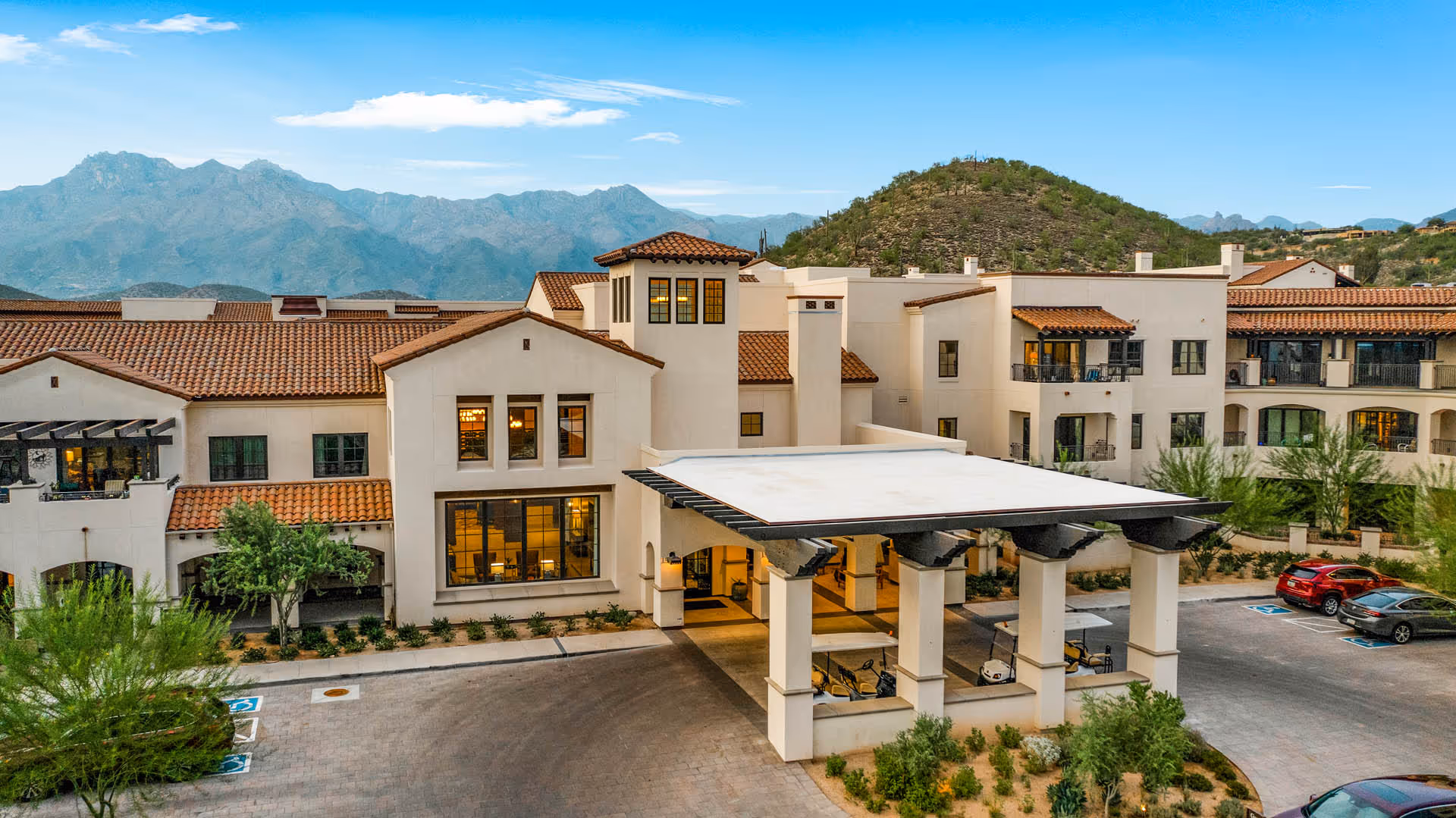 Exterior view of The Hacienda at the Canyon senior living facility with a covered entrance, multiple windows, balconies, and a parking area with cars. The building features a Spanish-style design with terracotta roof tiles and cream-colored walls, set against a backdrop of mountains and a clear blue sky.