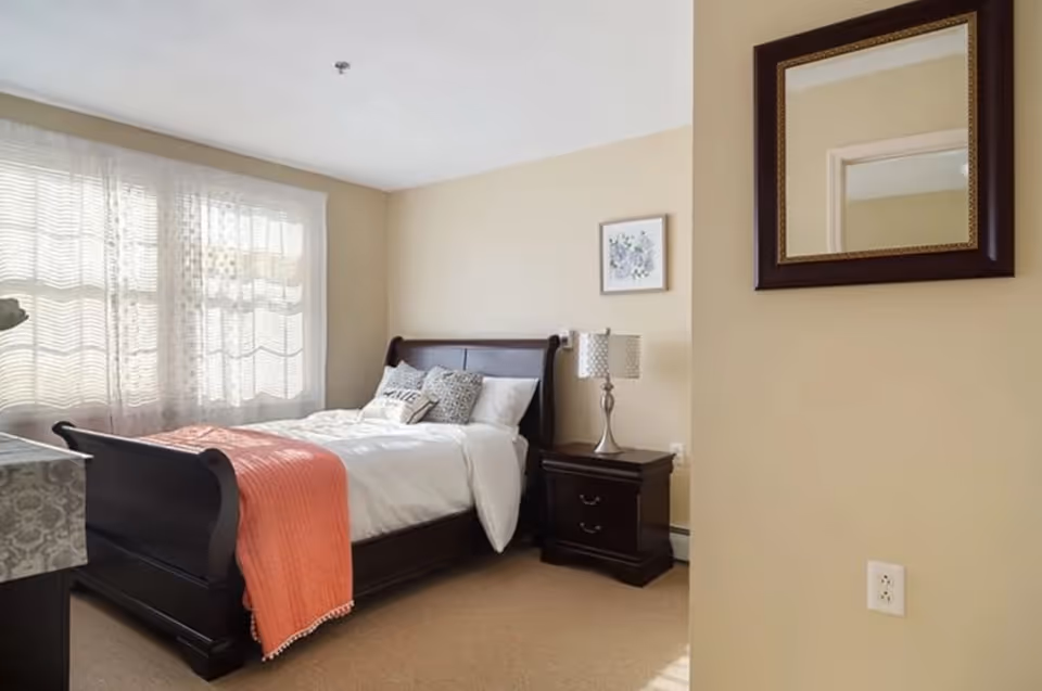 A cozy bedroom with a dark wooden sleigh bed made with white bedding and a coral throw blanket. There are decorative pillows on the bed, a nightstand with a lamp, a framed floral picture on the wall, and a large mirror hanging on the adjacent wall. Light filters through sheer curtains covering a window.