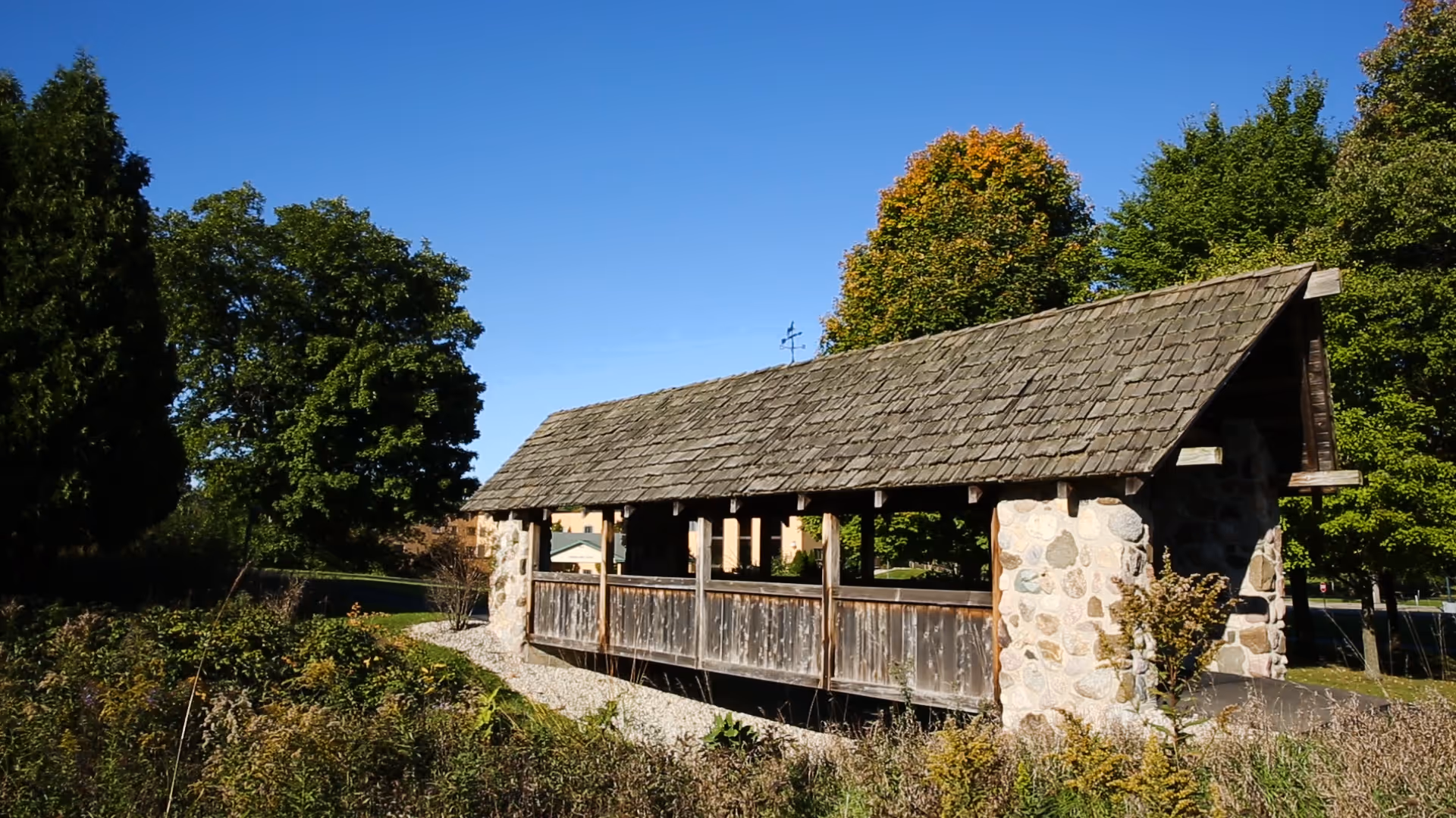 An outdoor scene featuring a long wooden covered structure with stone pillars and a shingled roof, set among trees under a clear blue sky.