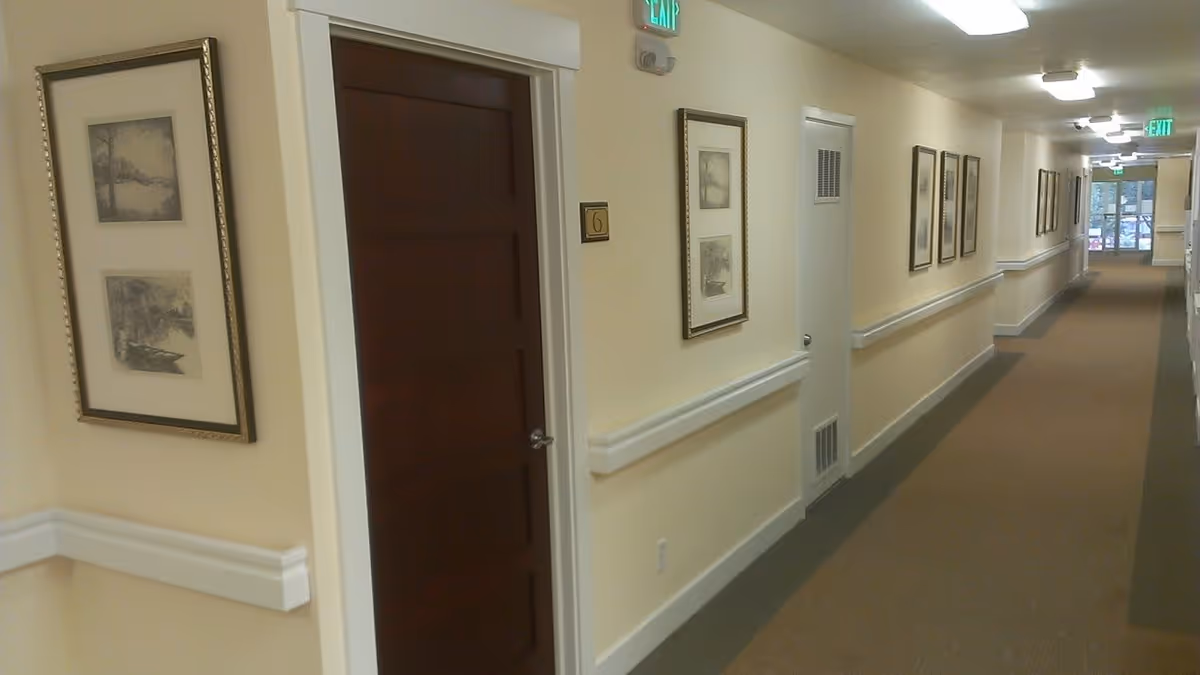 A long, well-lit hallway in a senior living facility with beige walls, brown carpet, framed artwork on the walls, and several doors including one labeled with the number 6. Handrails run along both sides of the hallway, and exit signs are visible at the far end.