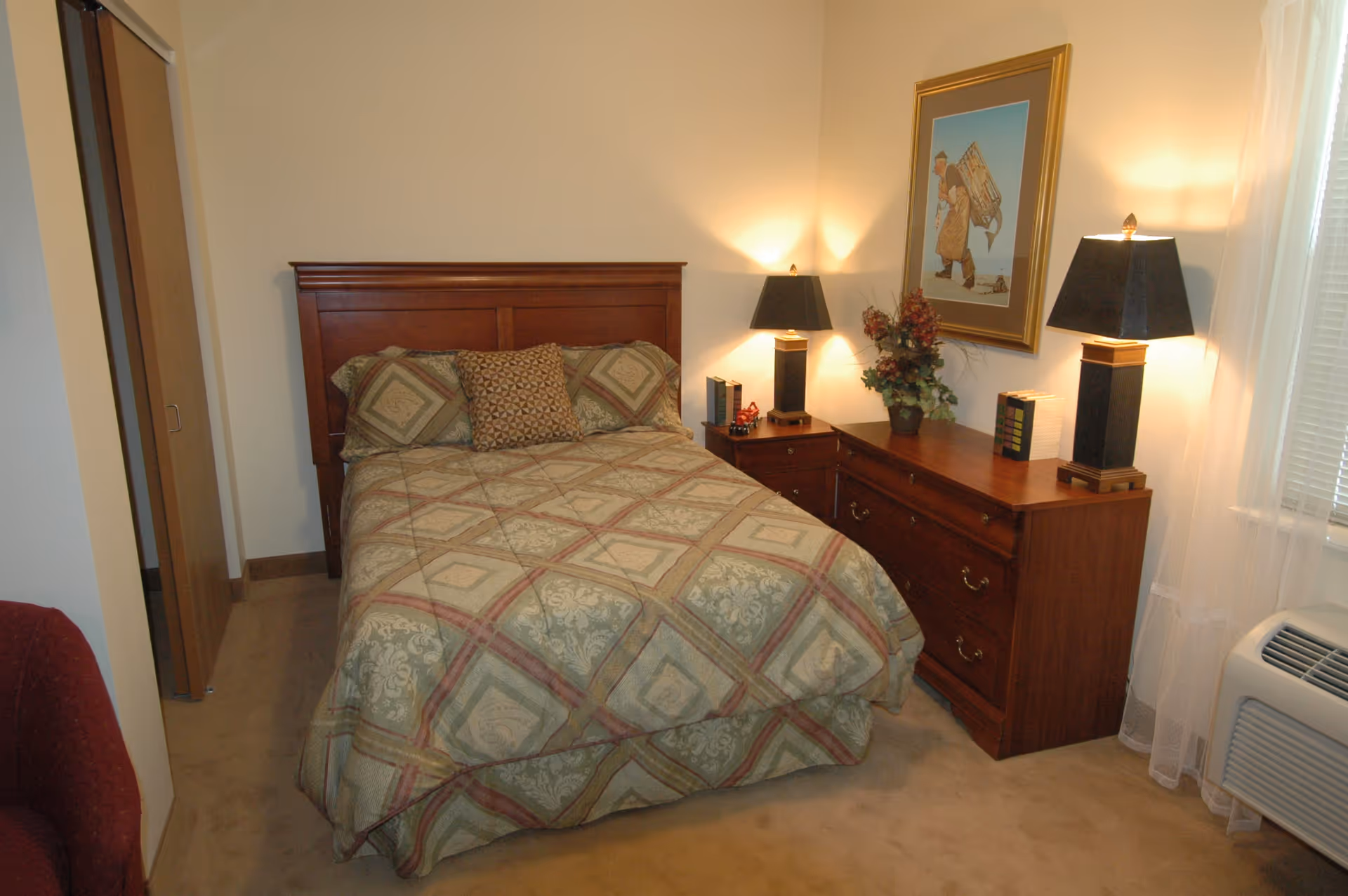 A bedroom with a wooden bed frame and a bed made with a patterned comforter and pillows. There is a wooden dresser with two black lamps, books, and a flower arrangement on top. A framed picture hangs on the wall above the dresser. A window with sheer curtains and an air conditioning unit is visible on the right side.