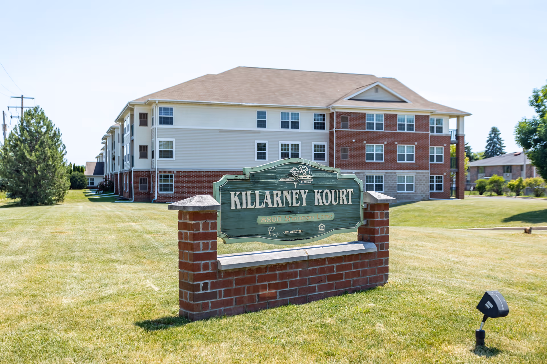A large three-story residential building with a mix of brick and siding exterior, surrounded by a well-maintained lawn and trees. In the foreground, there is a brick signpost with a green wooden sign that reads 'Killarney Kourt'.