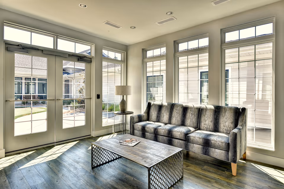 A bright living room area with large windows and glass doors letting in natural light. The room features a gray upholstered sofa, a wooden coffee table with a metal frame, and a side table with a lamp. Outside the windows, parts of the building and a small garden area are visible.