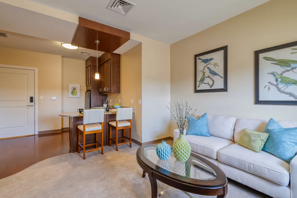 A cozy living area in a senior living facility featuring a beige sofa with blue and green cushions, a glass-top coffee table with decorative vases, and framed bird artwork on the wall. Adjacent to the living area is a small kitchen with a granite countertop, two cushioned bar stools, wooden cabinets, and pendant lighting.