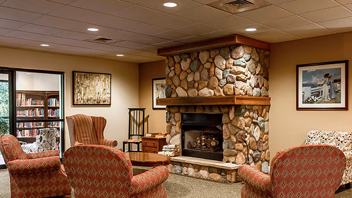 Cozy common area with a stone fireplace surrounded by four upholstered armchairs and a patterned sofa. The room has beige walls adorned with framed artwork, a wooden coffee table in the center, and a view into a library with bookshelves through a glass door.