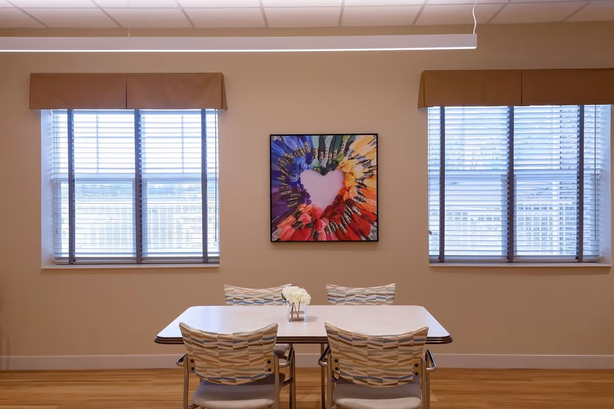 A small dining area with a rectangular table and four patterned chairs. The table has a small vase with white flowers. Behind the table are two windows with blinds and brown valances. A colorful framed artwork featuring embroidery floss arranged in a heart shape is hung on the wall between the windows.