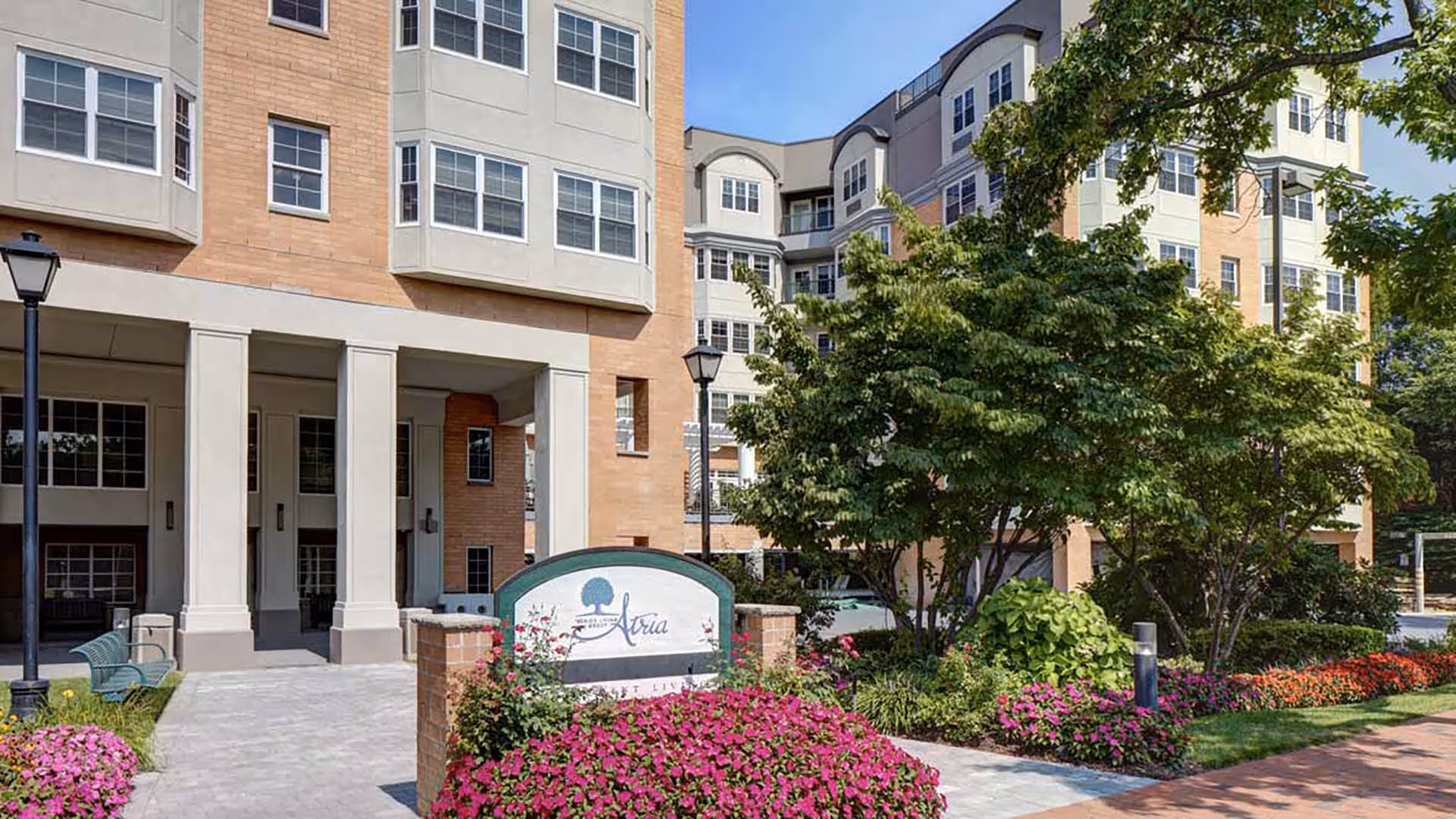 Exterior view of Atria Park of Great Neck senior living facility showing the building entrance with columns, landscaped garden with colorful flowers and trees, a sidewalk, and a sign with the facility name.