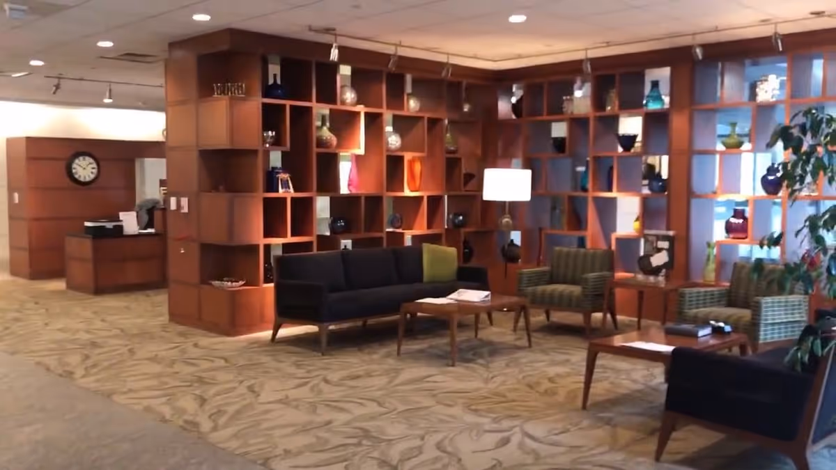 A cozy interior lounge area with a patterned carpet, wooden shelving filled with decorative vases, a black sofa, green patterned armchairs, wooden coffee tables, and a standing lamp. A reception desk and a wall clock are visible in the background.