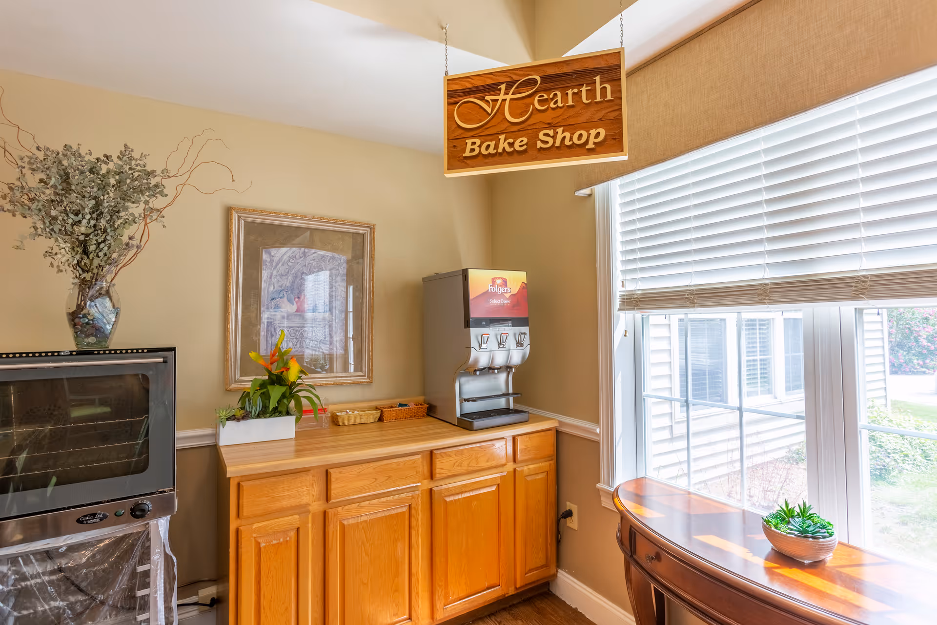 Interior view of a small bake shop area with a wooden cabinet, a coffee dispenser, a framed picture on the wall, and a window with blinds letting in natural light. There is a wooden sign hanging from the ceiling that reads 'Hearth Bake Shop'. A vase with decorative branches and a small plant are also visible.