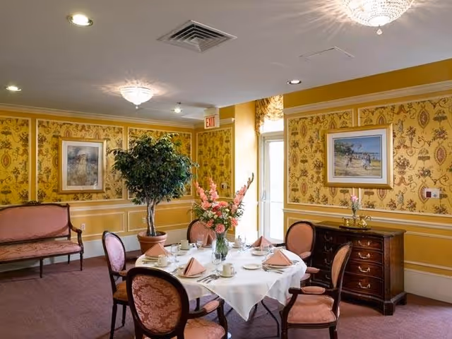 A dining room with a round table set for six people with pink napkins, white tablecloth, and floral centerpiece. The room has yellow patterned wallpaper, a potted tree, framed paintings on the walls, a wooden sideboard, and a pink upholstered bench. Ceiling lights illuminate the space.