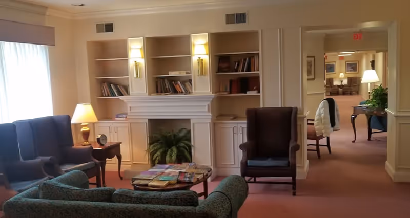 A cozy living room area in an assisted living facility featuring several upholstered chairs and sofas arranged around a coffee table with books and magazines. There is a fireplace with built-in shelves on either side, some containing books. A lamp on a side table provides warm lighting, and a hallway leads to another room with additional seating and lamps.