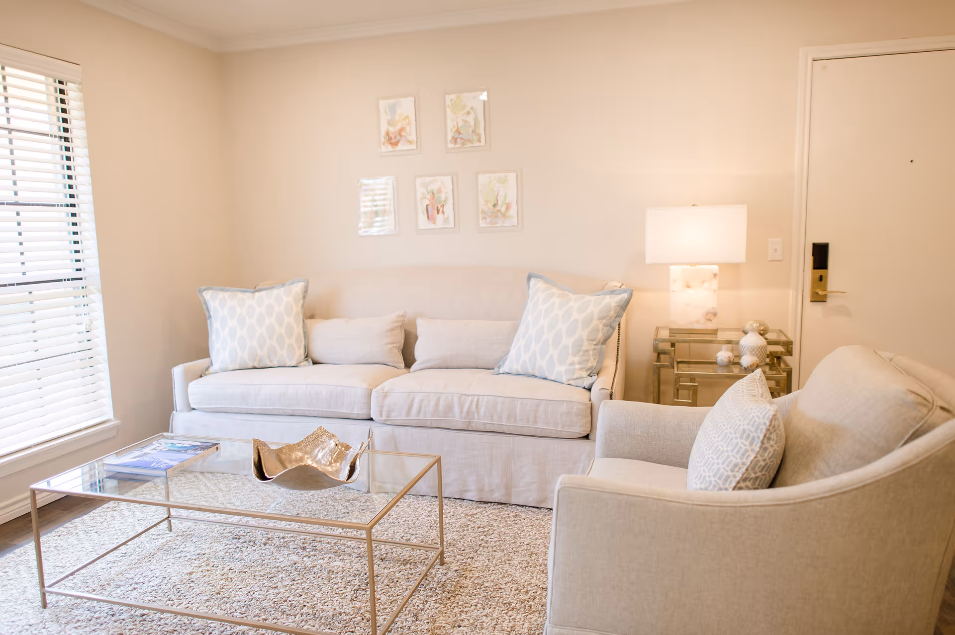 Bright living room with a beige sofa and armchair, glass coffee table, side table with lamp, and decorative pillows.
