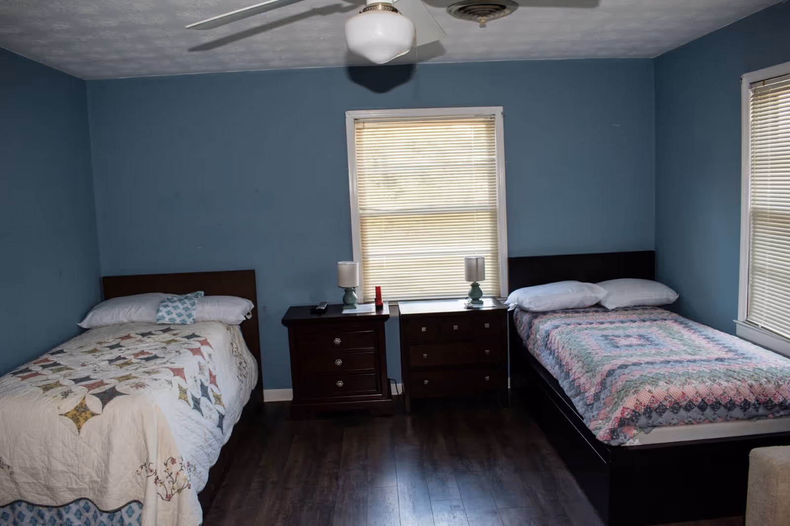 A bedroom with two single beds separated by two dark wooden nightstands. Each nightstand has a lamp on it. The walls are painted blue and there are two windows with closed blinds. The floor is dark wood and there is a ceiling fan with a light fixture above.