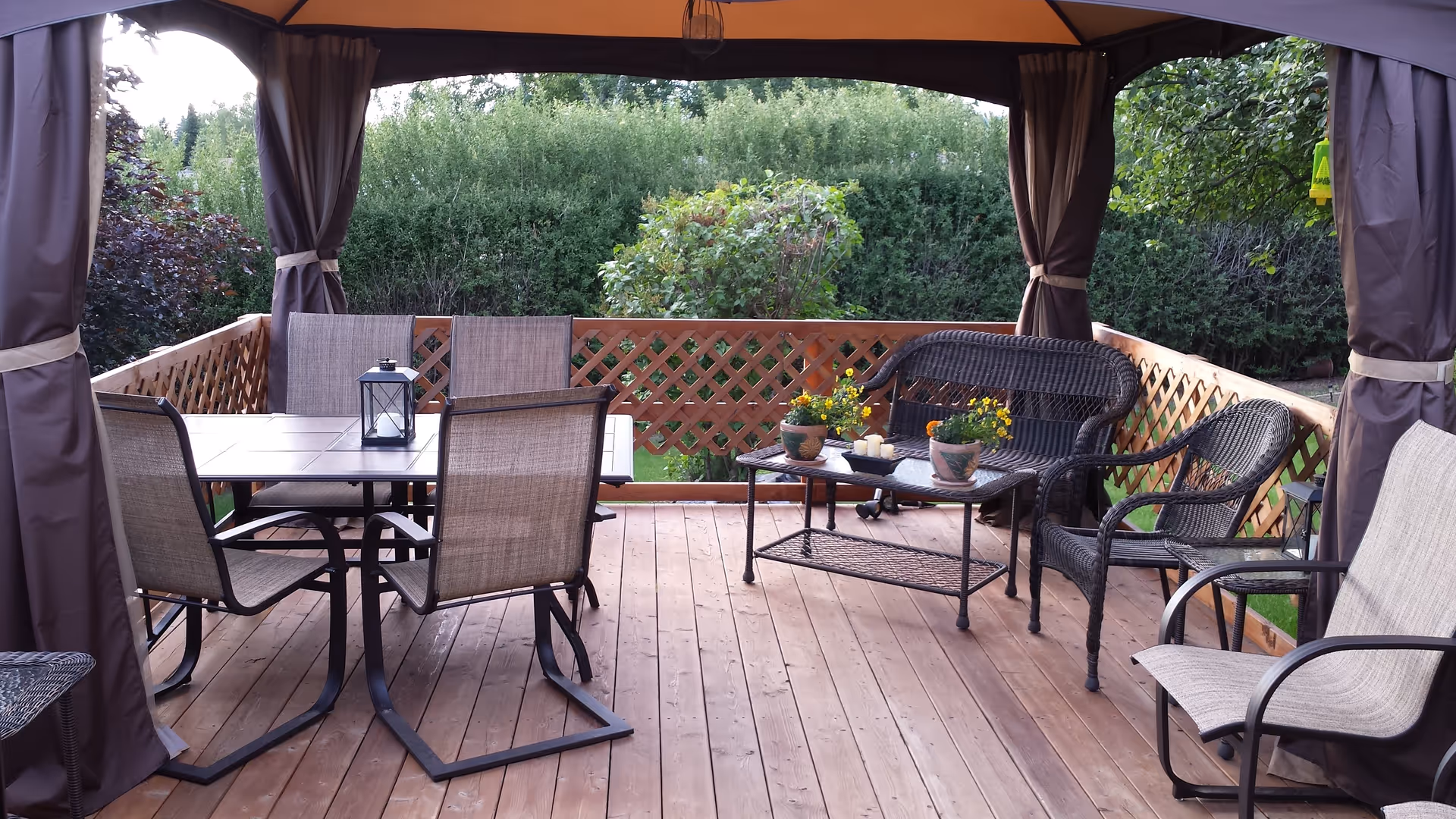 Outdoor covered patio area with wooden floor, featuring a dining table with four chairs, a small coffee table with potted flowers and candles, a wicker loveseat, and two wicker chairs. The patio is surrounded by a wooden lattice railing and green bushes in the background.