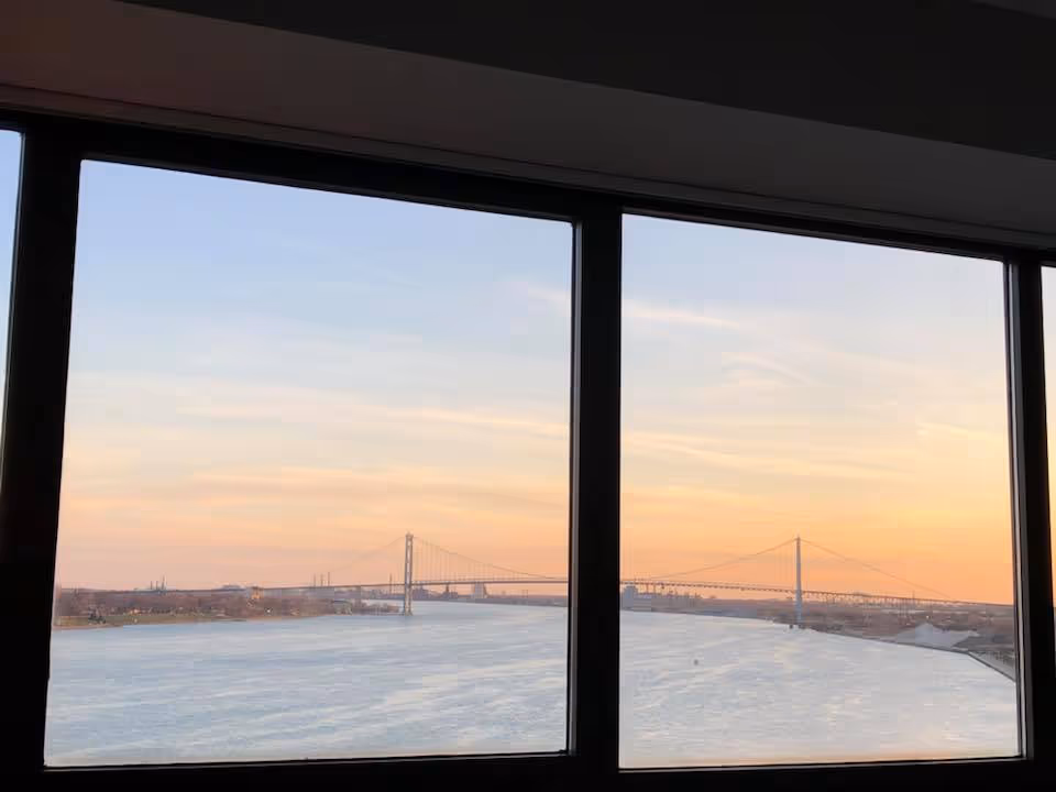 View of a river and a suspension bridge at sunset, seen through three large rectangular windows from inside a building.