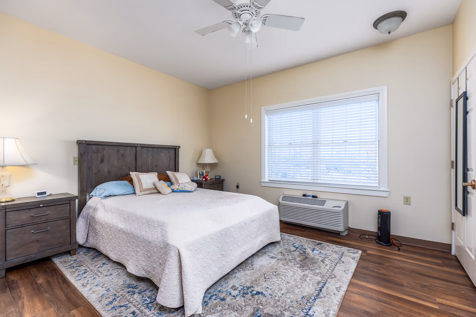 Bright bedroom with a bed, two nightstands, ceiling fan, window with blinds and wood-look flooring.
