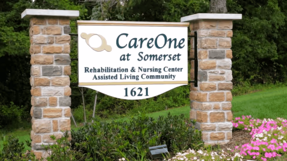 Stone pillars supporting a sign for CareOne at Somerset, a Rehabilitation & Nursing Center and Assisted Living Community, with the number 1621 below. The sign is surrounded by greenery and flowers.