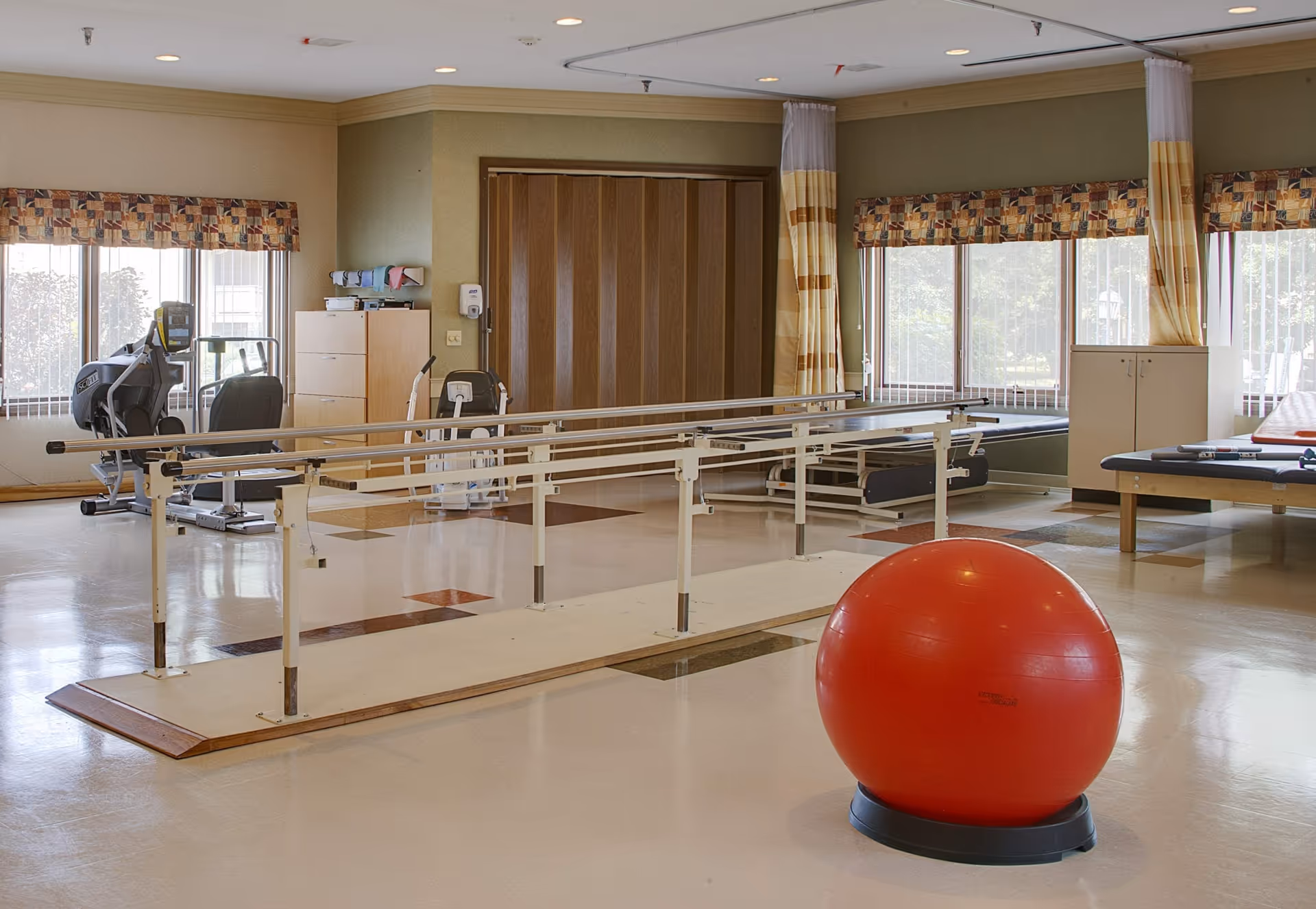 A rehabilitation therapy room with parallel bars for walking exercises, a large red exercise ball on a stand, exercise machines near windows with patterned valances, and therapy tables along the walls.