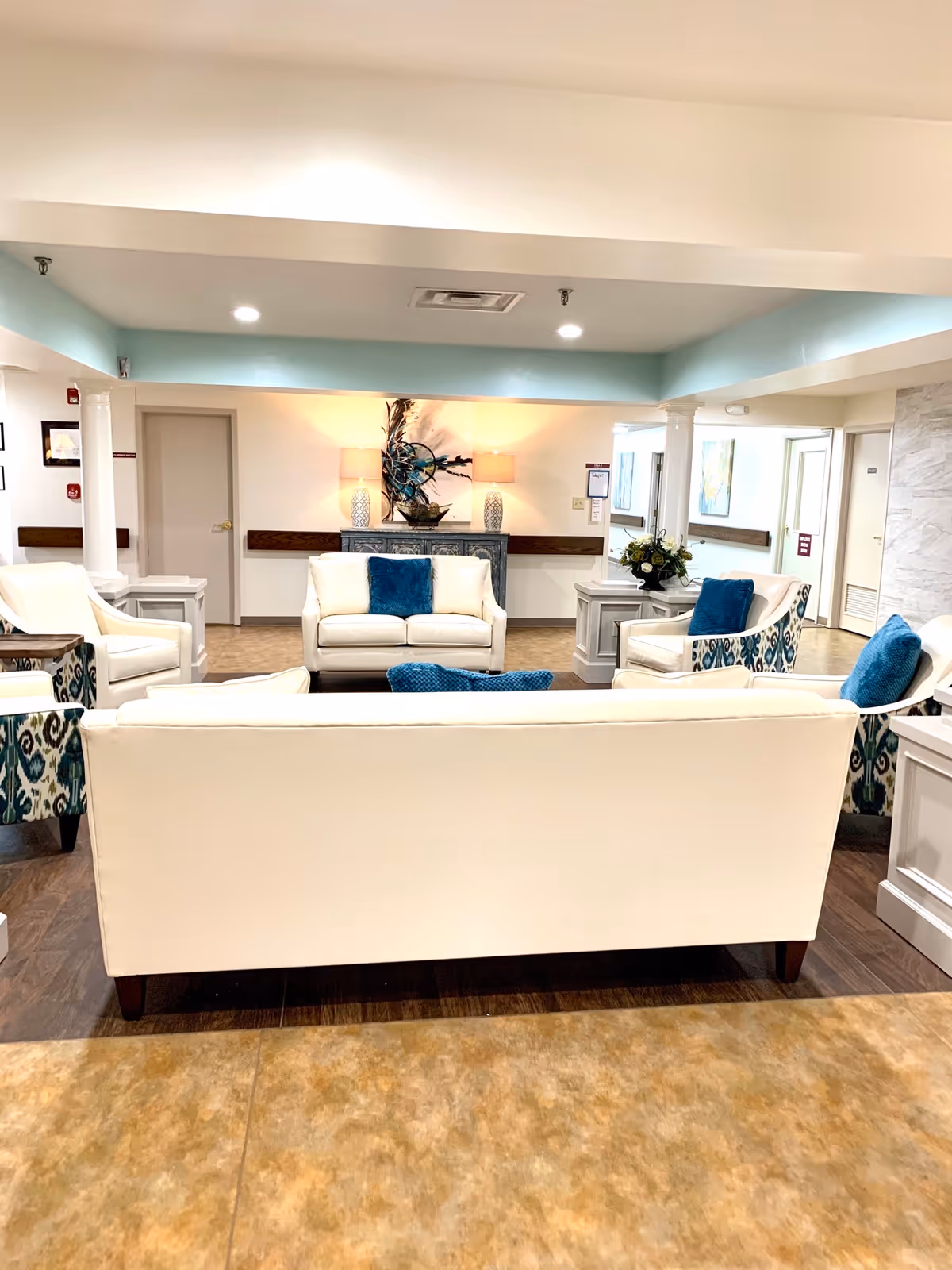 Bright communal lounge with white sofas, patterned armchairs and blue accent pillows facing a decorative console table and lamps.