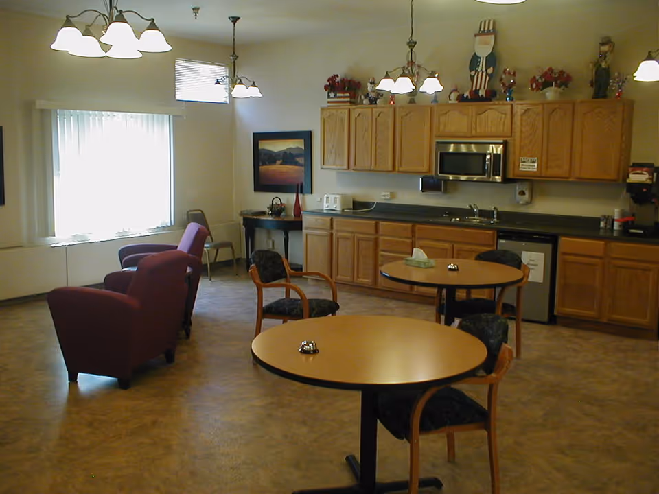 A communal room with a kitchenette featuring wooden cabinets, a microwave, a sink, and a small refrigerator. There are two round tables with chairs around them, a few armchairs near a window with vertical blinds, and decorative items on top of the cabinets including patriotic-themed figurines. The room is well-lit with ceiling lights.