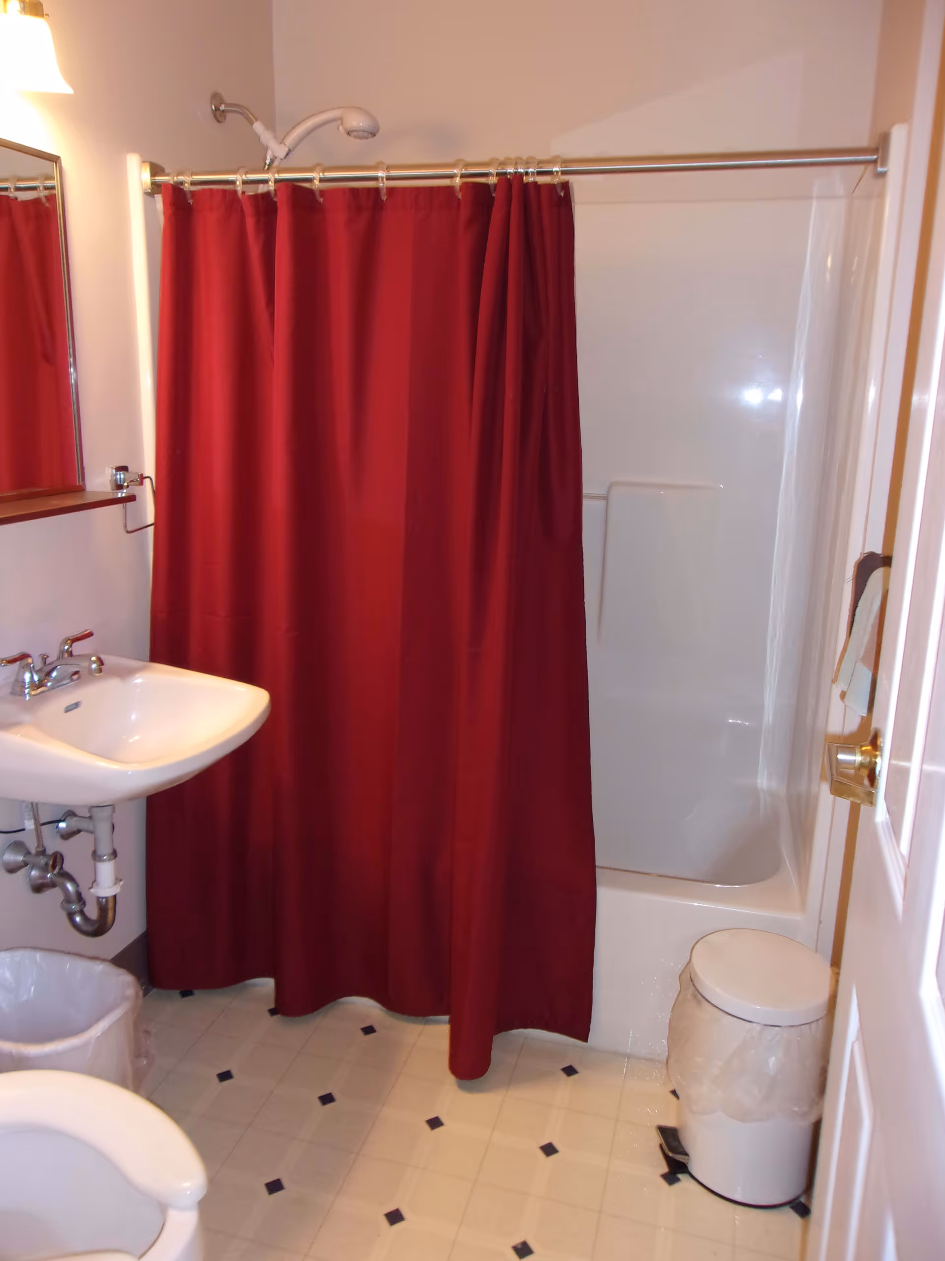 A small bathroom with a white sink, a toilet, a trash can, and a bathtub with a red shower curtain. The floor has a white tile pattern with small black diamonds. A towel hangs on the door.