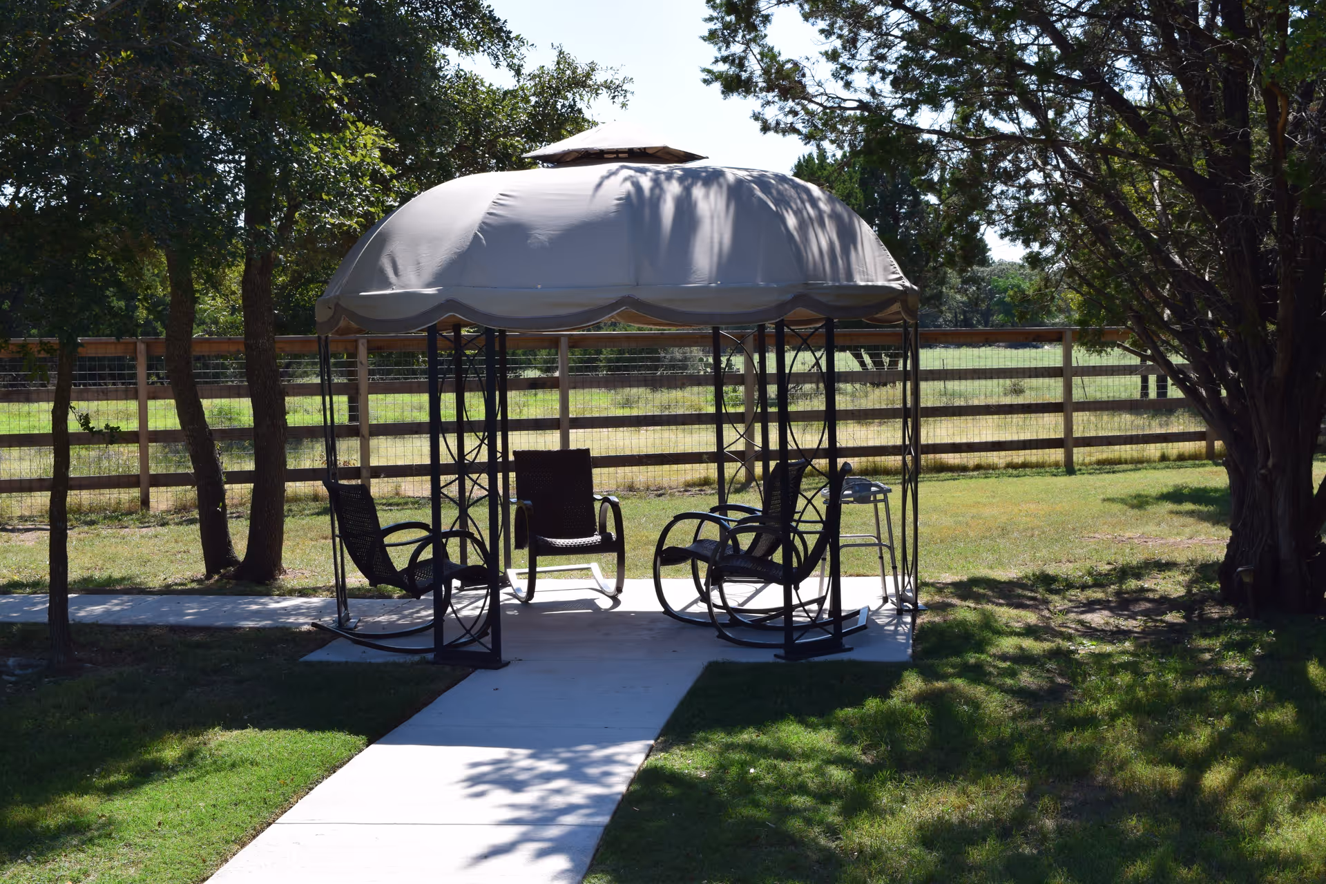 Outdoor gazebo with a beige canopy and four black rocking chairs arranged inside on a concrete slab, surrounded by green grass, trees, and a wooden fence in the background.