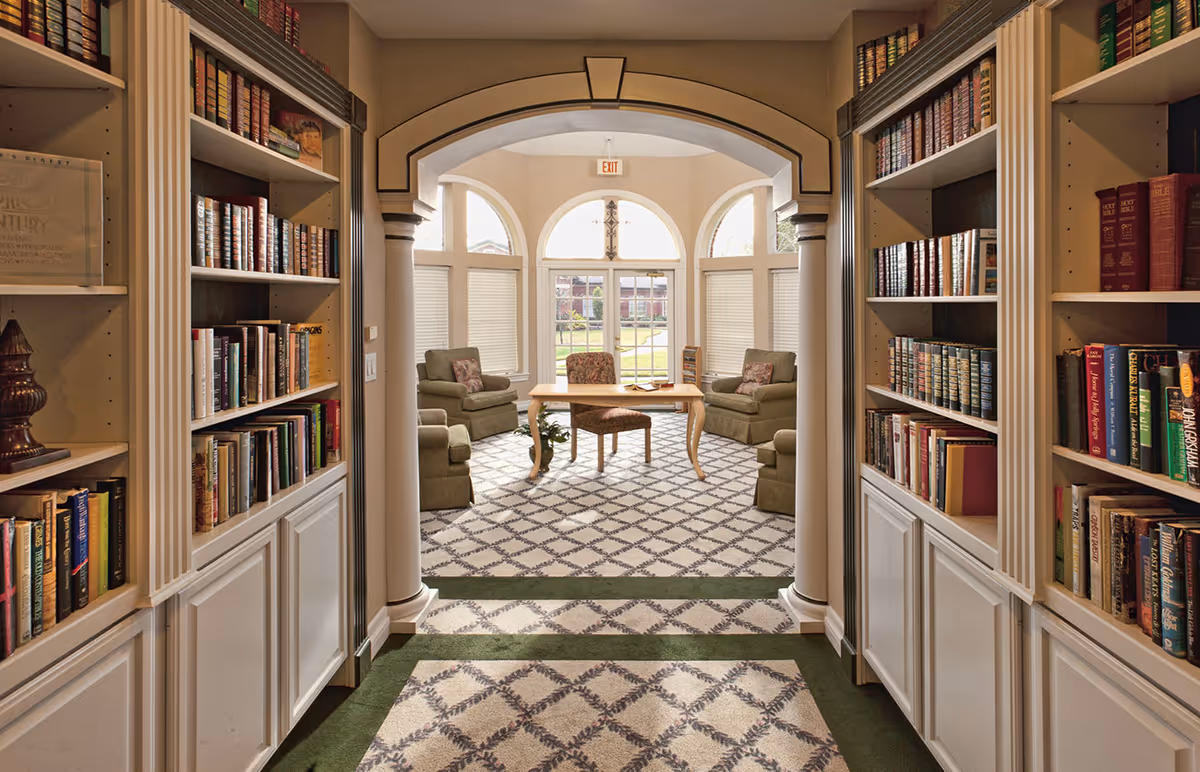 A cozy library room with built-in bookshelves filled with books on both sides of a hallway leading to a sitting area. The sitting area has a wooden table surrounded by four cushioned armchairs, with large windows letting in natural light and offering a view of the outdoors.